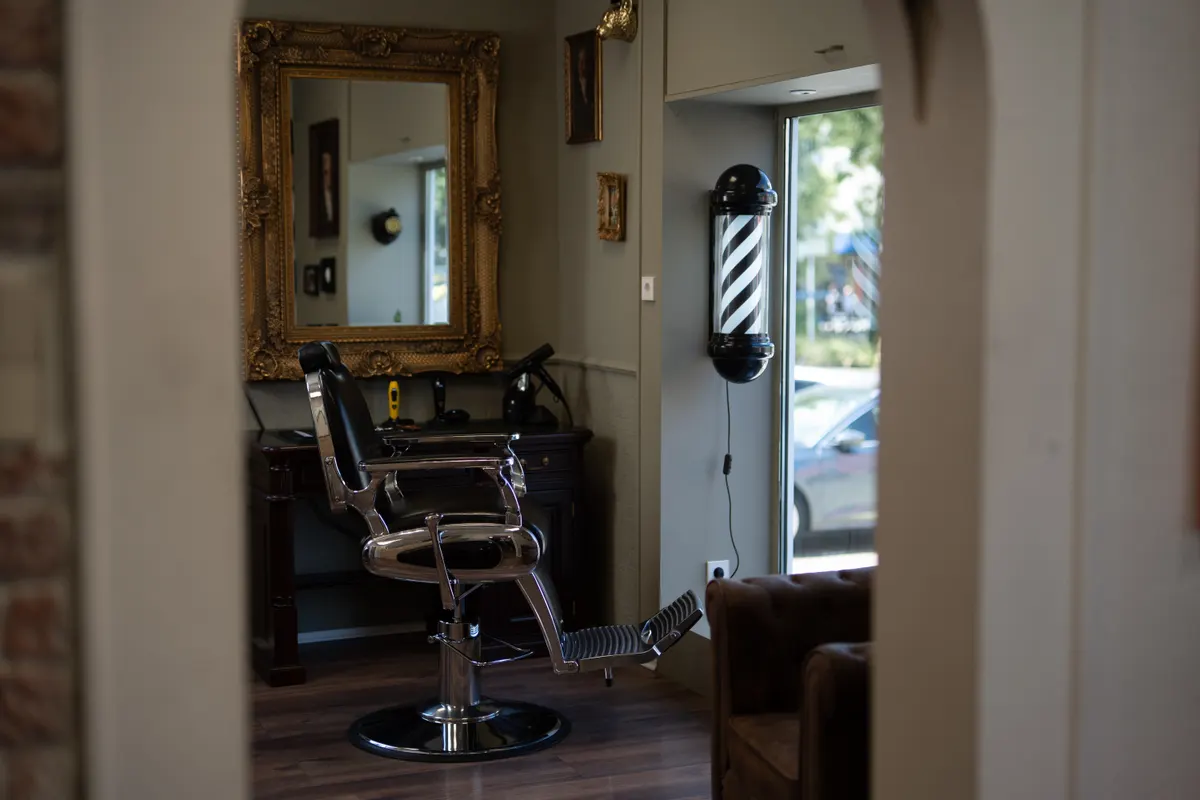 Blick in den Salon von Der Barbier: Barber-Stuhl vor goldgerahmtem Spiegel, Barber-Pole am Fenster