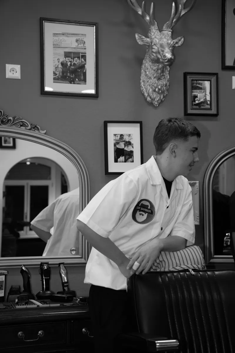 Len, Auszubildender, stützt sich im weißen Barbier-Hemd auf die Lehne des Barber-Stuhls